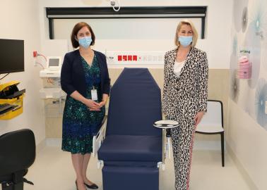 Two women stand either side of an examination chair in a clinical room. They are wearing masks.